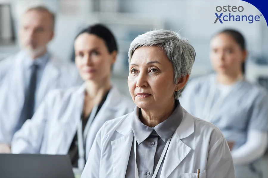 Eine Gruppe von Ärztinnen sitzt in einem Seminarraum. Im Fokus und Vordergrund ist eine ältere Medizinieren mit kurzen grauen Haaren. Jüngere Ärzte sind leicht verschwommen im Hintergrund.