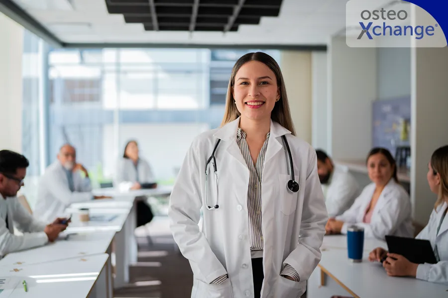 Eine Ärztin steht in einem mittelgroßen Meetingraum und schaut direkt aus dem Bild heraus. Im Hintergrund sitzen weitere Medizinier an den Tischen.