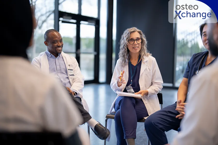 Eine Gruppe von Ärztinnen sitzt in einer Art Stuhlkreis und diskutiert. Im Vordergrund sieht man nur den Rücken einzelner Teilnehmer. Der Fokus liegt auf einer Frau in der Bitte des Bildes, die einen Notizblock auf dem Schoß hat.